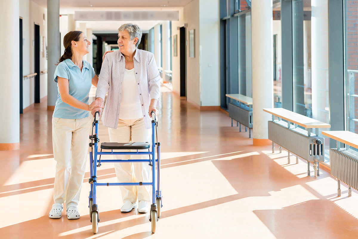 Frau geht an Rollator von einer Therapeutin unterstützt.