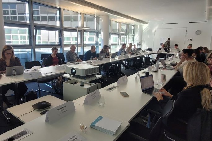 Blick in einen Konferenzraum, in dem Personen an einem U-förmigen Tisch sitzen.