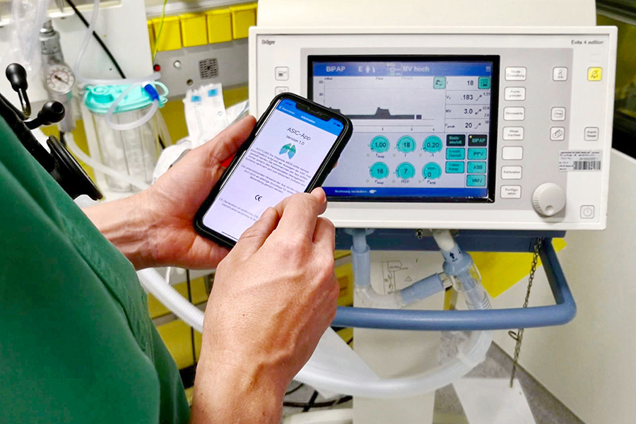 Ein Mitarbeiter auf der Intensivstation hält ein Mobiltelefon in den Händen und vergleicht die Daten einer medizinischen App mit einem Überwachungsgerät. 