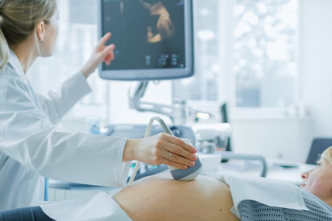 Bildbeschreibung: Eine schwangere Frau liegt mit dem Rücken auf einer Liege. Eine Ärztin im weißen Kittel hält ein Ultraschallgerät in der Hand und tastet den hochgewölbten Bauch der Schwangeren ab. Beide schauen auf einen Monitor.