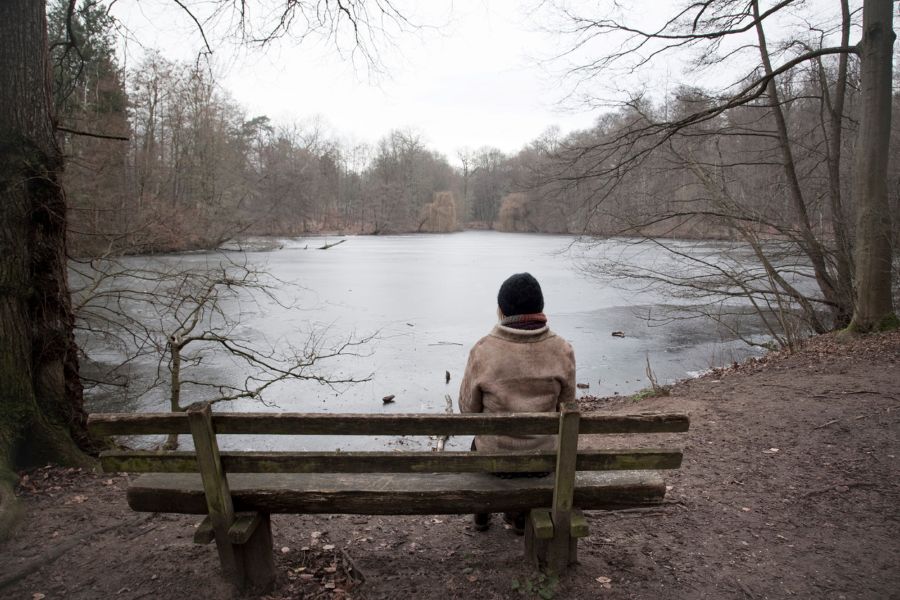 Eine Person sitzt an einem grauen Tag mit dem Rücken zum Betrachter auf einer Bank und schaut auf einen See. Eine Person sitzt an einem grauen Tag mit dem Rücken zum Betrachter auf einer Bank und schaut auf einen See.