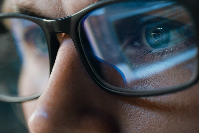Nahaufnahme einer Person mit Brille, in deren Gläsern der blaue Schein eines Computerbildschirms reflektiert wird