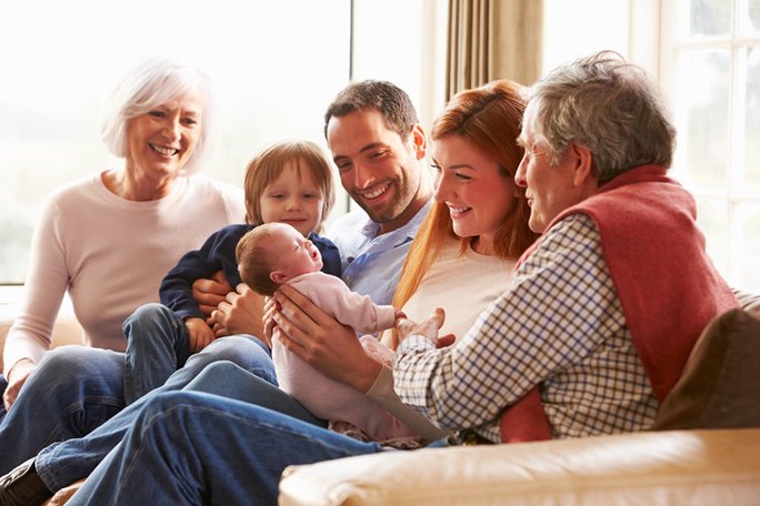 Eine Familie mit Großeltern, Eltern und Kindern sitzt auf einem Sofa und schaut auf einen Säugling