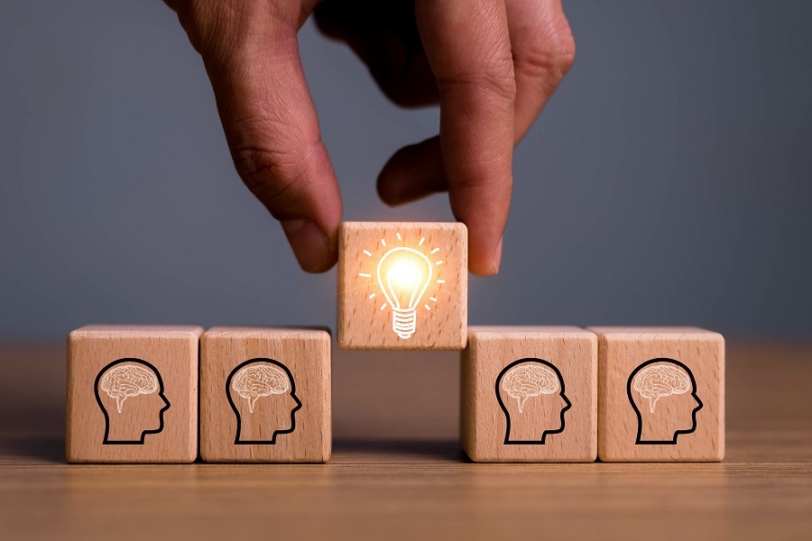 Symbolbild Kreativität, eine Hand stellt ein Holzklötzchen mit einer Glühlampe zwischen Holzklötzchen mit einem Kopf