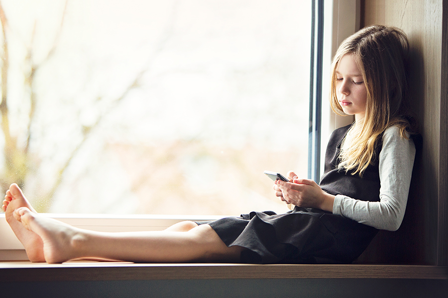 Kleines Mädchen sitzt auf der Fensterbank und spielt mit dem Smartphone.