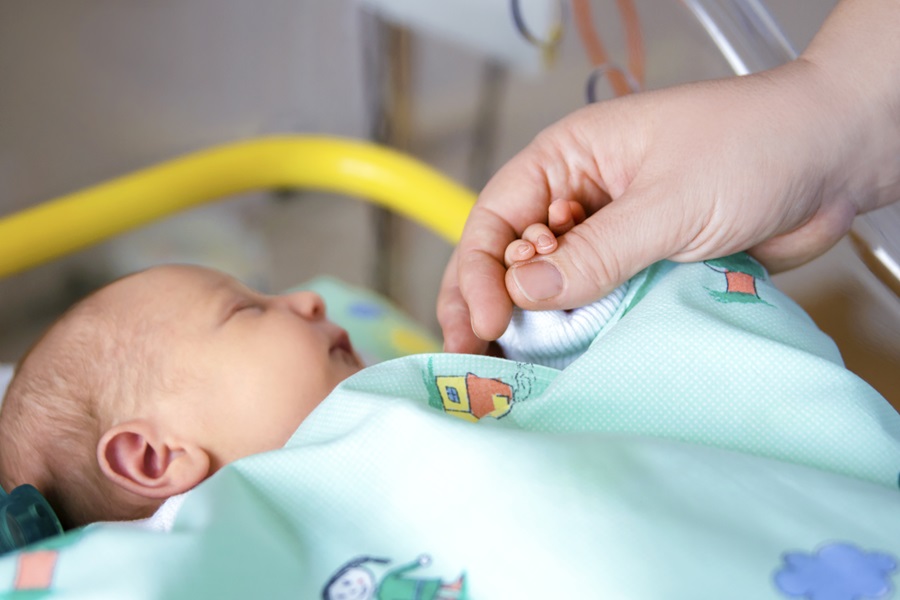 Ein S&auml;ugling liegt schlafend in einem Bett, eine Hand wird von der Hand eines Erwachsenen gehalten