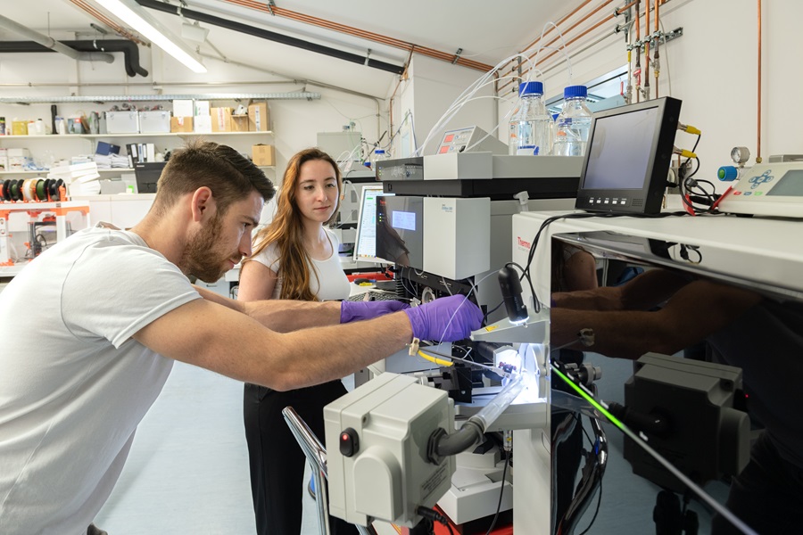 Ein junger Mann arbeitet an einem Massenspektrometer, seitlich steht eine junge Frau