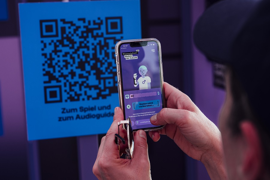 Bei der Ausstellung Health XP h&auml;lt eine Person ein Smartphone, auf dem ein Avatar zu sehen ist, vor einen QR Code