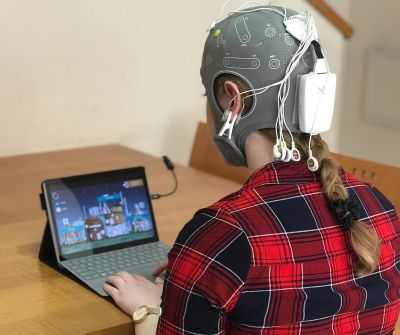 Eine Probandin sitzt an einem Laptop und spielt ein Videospiel. Auf dem Kopf trägt sie Elektroden zur Hirnstimulation. Eine Probandin sitzt an einem Laptop und spielt ein Videospiel. Auf dem Kopf trägt sie Elektroden zur Hirnstimulation.
