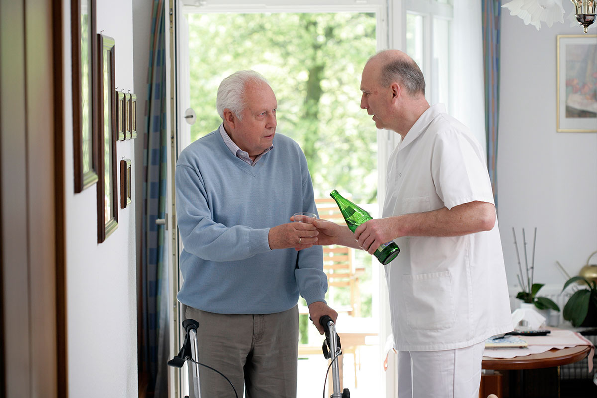 Ein Arzt begrüßt einen älteren Patienten mit Rollator an der Eingangstür der Praxis.