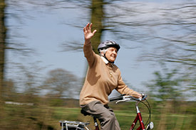 Eine ältere Dame mit Fahrradhelm fährt auf einem Fahrrad und winkt