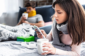Im Vordergrund kranke junge Frau mit einer Tasse Tee und einem Smartphone in der Hand