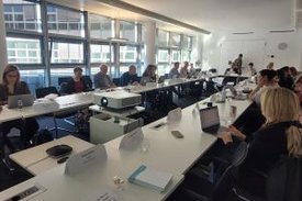 Blick in einen Konferenzraum, in dem Personen an einem U-förmigen Tisch sitzen.
