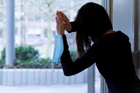 Frau mit Maske in der Hand lehnt sich an eine Fensterscheibe