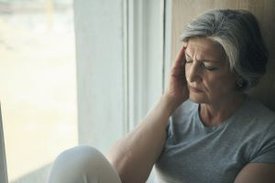 Eine Frau sitzt mit geschlossenen Augen an einem Fenster und hält sich mit der rechten Hand die Stirn.