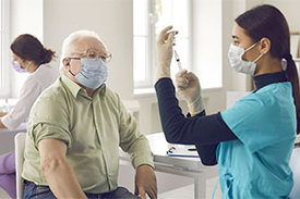 Medizinische Fachangestellte zieht Spritze mit Impfstoff auf, um vor ihr sitzenden älteren Mann zu impfen