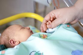 Ein Säugling liegt schlafend in einem Bett, eine Hand wird von der Hand eines Erwachsenen gehalten