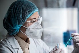 Frau in Laborkleidung mit Maske untersucht Flüssigkeit in einem Reagenzglas.