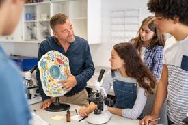 Lehrer erklärt vier Jugendlichen an einem überlebensgroßen Modell die Funktion einer Zelle, während eine der Schülerinnen vor einem Mikroskop sitzt. 