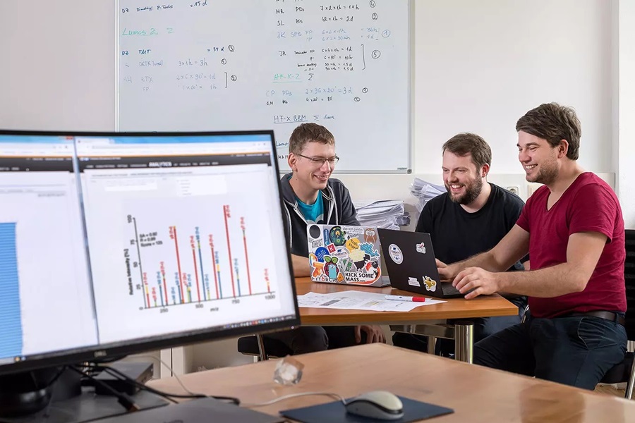 Im Vordergrund stehen zwei Bildschirme, im Hintergrund rechts sitzen drei junge Männer um Laptops